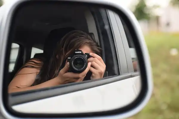 A young woman taking a picture of herelf in a car's side view mirror.